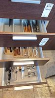 Three open drawers showing assorted wood-handled knives, flatware organiser with tools, lower drawer with utensils and jug