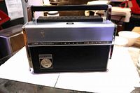 Front view of Zenith Trans-Oceanic radio showing brushed metal upper section, black lower panel, crest emblem, carry handle