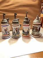 Front view of all four steins showing size, decoration, and lids