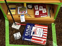 Shelf contents showing boxed flag, miniature Eiffel Tower, ring mandrel, screwdriver, playing card deck, binder, alarm clock, card games, writing instruments