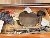Drawer view displaying grill pan, saucepan, gloves, utensils, skewers, lids and accessories