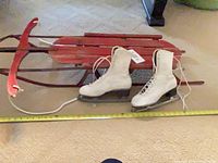 Side view of sled with metal runners, pair of white skates in front, tape for scale