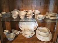 Photo showing a collection of various plates, teacups, and serving pieces arranged on two shelves. Porcelain pieces decorated with floral pattern. Includes stacked plates, cups, and platters.