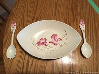 Overview photo of the white elongated oval dish with pink magnolia floral hand-painted inside and matching salad tongs on either side, shown on wooden table.