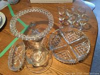 Various crystal and cut glass items displayed on wooden table including pedestal cake stand, sectional serving dish, candy dish, oval dish, and glass stir stick.