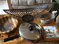 Table with Lagostina saute pan, metal fruit basket, wooden bowl, metal basket, wooden cutting board, ceramic tile, and dust bin.