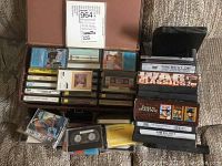 Photo showing boxes with DVDs, VHS tapes, and cassette tapes, with visible titles including Johnny Carson VHS, War Classics DVDs, and John Wayne DVD Collection.