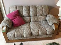 Photo showing the beige patterned upholstered love seat with wooden trim on the armrests and base, accompanied by a magenta pillow on the left side, placed against a wall on a carpeted floor.