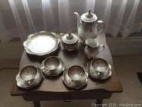 Full tea set displayed on wooden table in front of a window, showing cups, saucers, teapot, creamer, sugar bowl, and plates