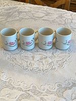 Four cream ceramic mugs with pink flower motif lined up on lace tablecloth