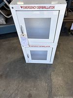 Front view of stacked white metal emergency defibrillator cabinets with windows and red lettering