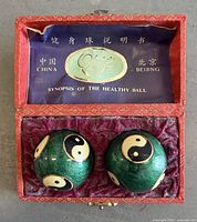 Open red brocade box showing two green yin-yang cloisonné Baoding balls seated on purple fabric, booklet affixed inside lid