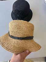 Both hats front view: straw hat held foreground, black felt cloche behind