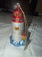 Front view of glass lighthouse ornament showing red roof, silver tower, blue base and gold cord