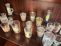 Group of souvenir shot glasses on shelf showing variety of prints, shapes and gold rim detailing.