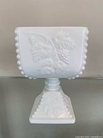 Front view of white milk glass pedestal compote showing grape relief and hobnail rim