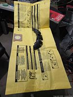 Yellow paper vacuum filter bag laid flat with printed instructions and cardboard collar, black plastic accessory holder placed on top