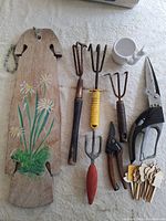 All gardening items laid out on white surface: wooden pot hanger, hand tools, ceramic planter, row markers