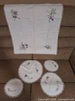 All five embroidered linens displayed: square cloth draped, four round doilies laid flat