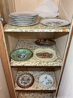 Overview of cupboard shelves showing multiple decorative plates including stacked gold-rimmed dinner plates, individual scenic and botanical plates