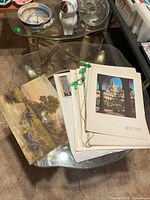 Stack of prints next to oil landscape painting on glass table