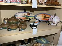 Shelf view showing stoneware teapot set, two landscape platters, porcelain cup and saucer, demitasse cups and saucers