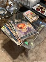 Stack of assorted vintage magazines on glass table, top issue The American Home, additional Mechanix Illustrated booklets visible