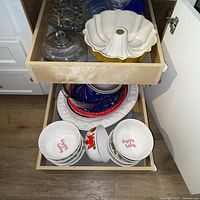 Two open drawers showing mixed dishware including bundt pan, glass dome and stand, bowls and plates