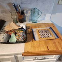 Wide view showing all items grouped on cutting board: wood board, plastic board, trivet, glass pitcher & vase, wire basket with contents