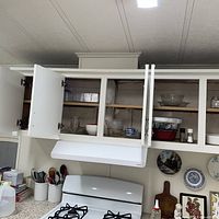Open upper cabinets above stove showing glass mixing bowl, ceramic bowls, spices, additional glassware