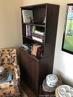 Photo of a tall brown wooden bookshelf filled partially with books on two open shelves and closed cabinet doors below, placed against a wall near a TV and a patterned armchair.