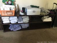 Brown wood TV stand with multiple shelves filled with fabric and some electronic items on top, positioned against a wall on carpeted floor.