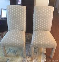 Two matching side chairs with reupholstered shiny stone color fabric featuring ivory detail pattern, placed on a rug in a living room.