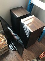 Two Polk Audio bookshelf speakers with wood veneer finish standing side by side, showing tops and sides. Positioned on wood floor with other items partially visible around.
