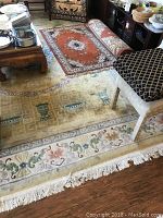 Photo of a large gold and green area rug featuring urn and floral motifs with a tan and orange small rug partially visible on top. The rug is laid on wooden flooring and there is a chair positioned on the rug.