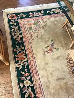 Partial view of the tan, green, and burgundy floral patterned rug showing the decorative border and some central motifs, illustrating condition and minor wear.