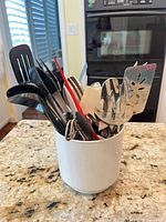 White ceramic crock holding assorted stainless steel and nylon cooking utensils on granite countertop