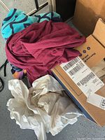 Mixed linens including teal-black striped towel, maroon towel, off-white sheet, cardboard box