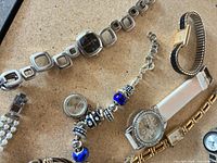 Overview showing square-link watch, charm bracelet with blue beads, white strap chronograph watch, gold-tone link watch, pearl strap watch, gold/black expansion watch