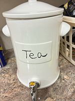 Front view of white ceramic beverage dispenser showing lid, side handles, label area, and spigot