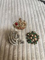 Three brooches laid out: gold crown, silver bird, green floral