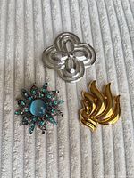 Group shot showing all three brooches: silver filigree with blue stones, gold flame motif, silver rosette