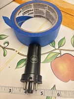 Side view of black 6V6 vacuum tube next to ruler showing length, supported by blue tape ring