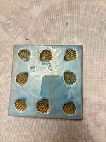 Front view of square ceramic coaster showing light blue glaze and eight tan shell motifs