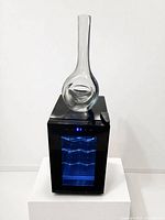 Full view of glass decanter resting on top of black wine cooler with blue interior light