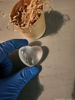 gloved hand holding clear heart crystal beside open cylindrical box with straw filler