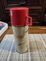 Thermos front showing red lid, beige woodgrain body, THERMOS logo and fly graphic