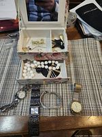 Open floral jewelry box showing contents: earrings, pins, necklace, watches, bracelet