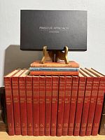 Front view of entire lot showing 15 red encyclopedia volumes, three vintage textbooks stacked on top, Pimsleur audio box on stand