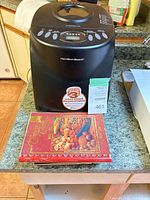 Front view of Hamilton Beach bread maker next to spiral-bound cookbook